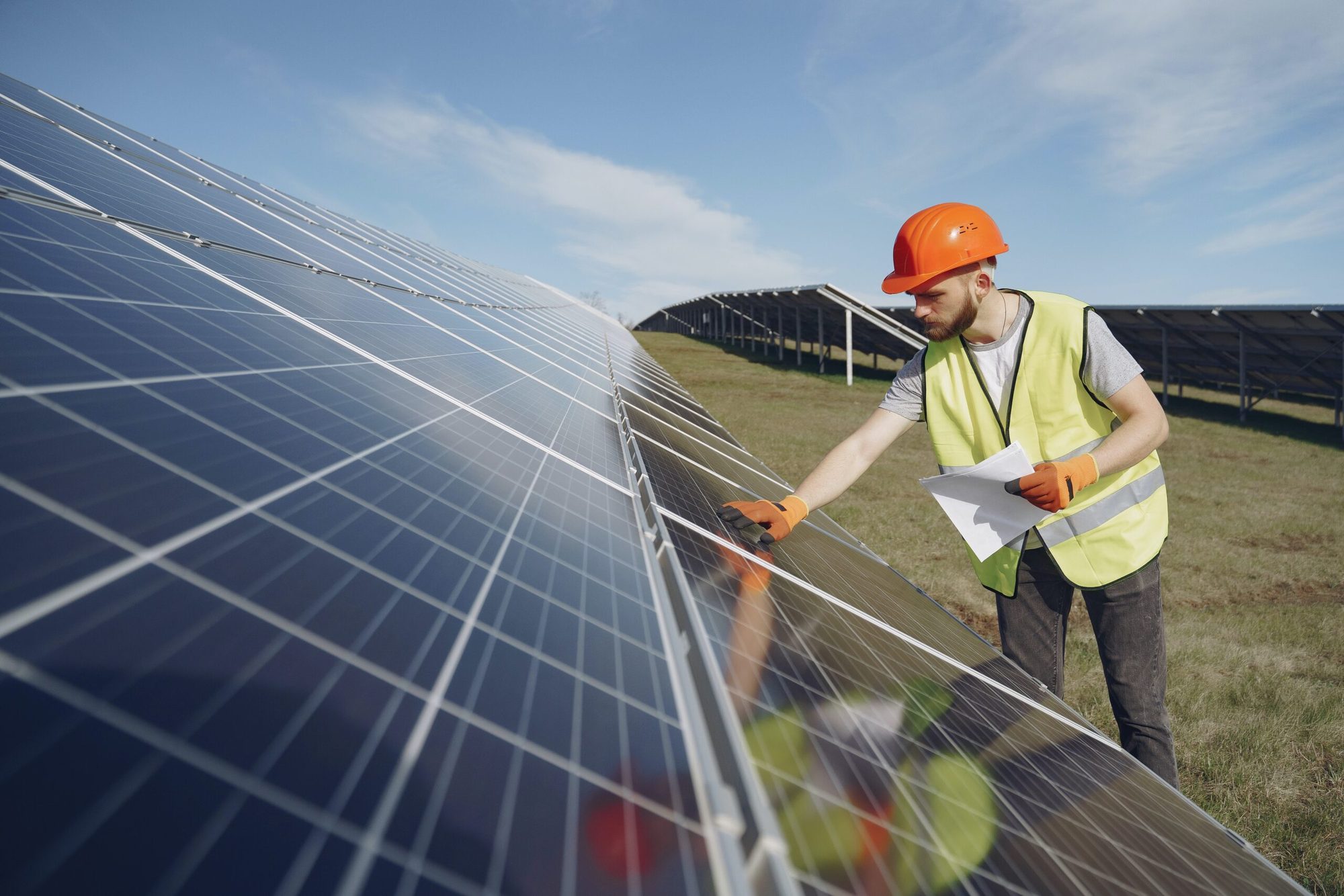 Techniker mit Schutzhelm und Warnweste prüft die Modulreihen eines Solarparks